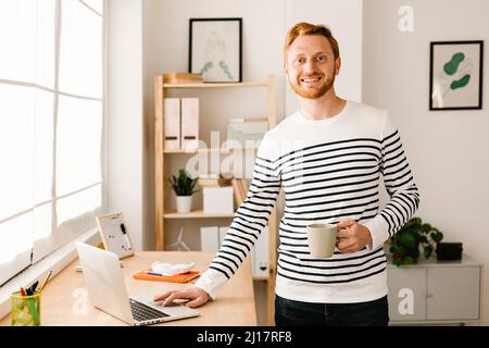 Sorridente uomo che tiene la tazza di caffè in piedi accanto al portatile a casa Foto Stock