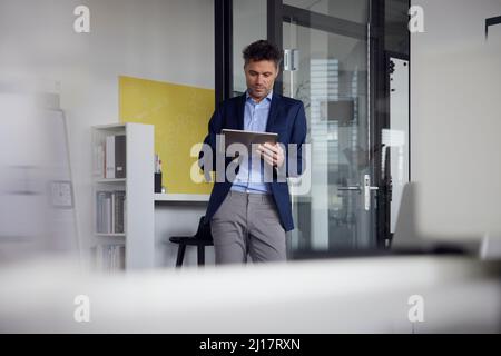 Uomo d'affari che utilizza un tablet PC appoggiato su una porta di vetro sul posto di lavoro Foto Stock