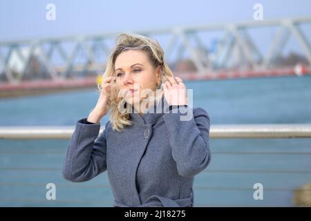 Donna attraente in piedi su una passeggiata lungomare nel vento che si aggrappa i suoi lunghi capelli biondi con le mani Foto Stock