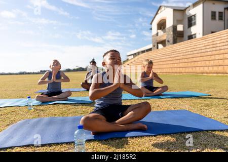 Ragazzi di scuola elementare multirazziale seduti su materassino da ginnastica mentre meditano in campo scolastico Foto Stock