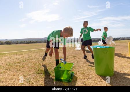 Ragazzi di scuola elementare multirazziale che mettono rifiuti di plastica nel cestino della spazzatura sul terreno della scuola Foto Stock