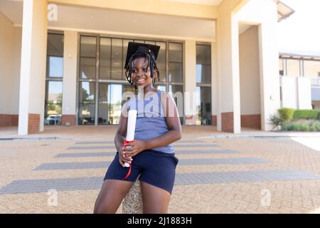 Scuola elementare afroamericana con mortarboard che tiene la laurea mentre si siede contro la scuola Foto Stock