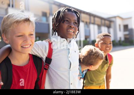 Sorridenti ragazzi di scuola elementare multirazziale con braccio intorno in piedi al campus durante la giornata di sole Foto Stock