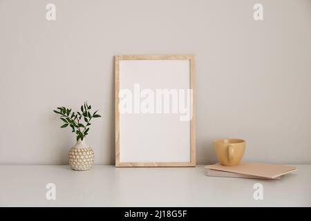 Immagine verticale del fotogramma su un tavolo bianco. Vaso con ramo verde, tazza di tè, taccuino di carta. Sfondo bianco della parete. Soggiorno in stile nordico Foto Stock