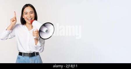 Sorridendo ragazza asiatica felice che parla in megaphone e che punta in su, annunciando promo di sconto, mostrando la pubblicità in cima, in piedi su sfondo bianco Foto Stock