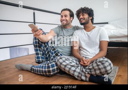 Coppia gay gioiosa seduta sul tappetino durante il channel-surf Foto Stock