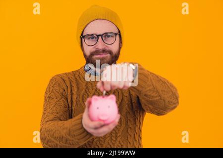 Giovane uomo caucasico bearded in cappello e occhiali che mettono le monete nel risparmio della banca del piggy e deposito concetto arancione sfondo isolato studio ritratto girato . Foto di alta qualità Foto Stock