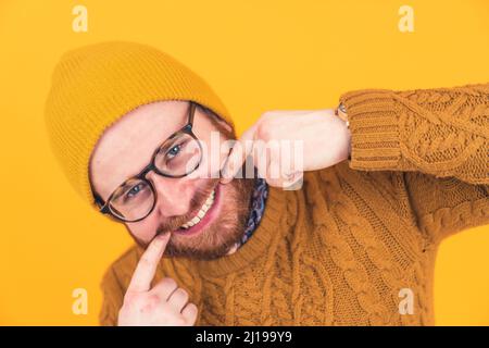 Ritratto del giovane caucasico sorridente in occhiali, cappello e barba, uomo che tocca i suoi bordi della bocca, mostrando denti arancione sfondo isolato studio shot . Foto di alta qualità Foto Stock