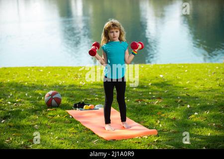 Carino bambino ragazzo pompando i muscoli del braccio con dumbbell all'aperto nel parco estivo. Fitness bambini con manubri. Foto Stock