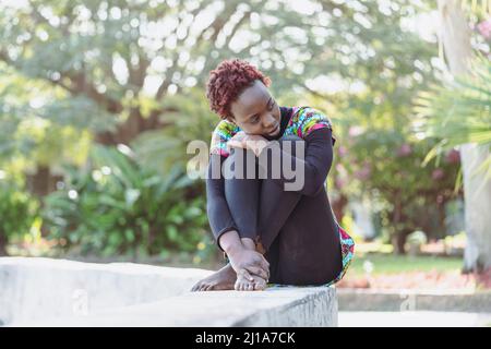 Giovane ragazza africana malinconica, seduta su un muro con le braccia intorno alle ginocchia, guardando contemplativamente lateralmente; affrontare i problemi e di Foto Stock