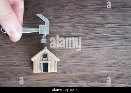 Dita che tengono un martello in acciaio miniaturizzato su un minuscolo modello di casa giocattolo su uno sfondo di legno. Progetto di ristrutturazione della casa. Foto Stock