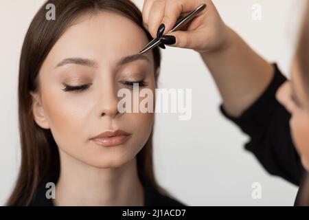 Primo piano di una giovane donna perfetta con capelli lunghi e scuri che pinzano le sopracciglia di un make-up artista professionista in abiti neri. Foto Stock