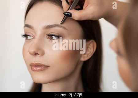 Primo piano di giovane donna splendida con lunghi capelli scuri che pinzano le sopracciglia da professionista make-up artista in abiti neri. Foto Stock