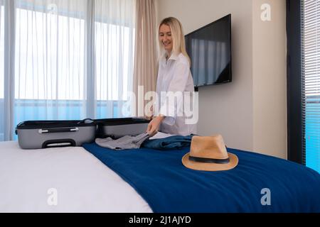 Allegra giovane donna felice in una camicia bianca raccoglie o scompatta le cose da una valigia che si trova sul letto. Concetto di viaggio, vacanza Foto Stock