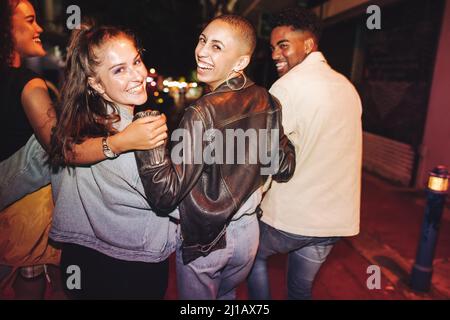 Buon tempo il sabato sera. Vista posteriore di un gruppo di amici vibranti ridere felicemente mentre camminano in città di notte. Patatine fritte multiculturali Foto Stock