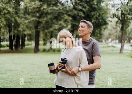 Una coppia matura e felice che ama abbracciarsi all'aria aperta nel parco cittadino. Un biondo caucasico uomo e donna passare il tempo insieme e bere caffè. Senior Foto Stock