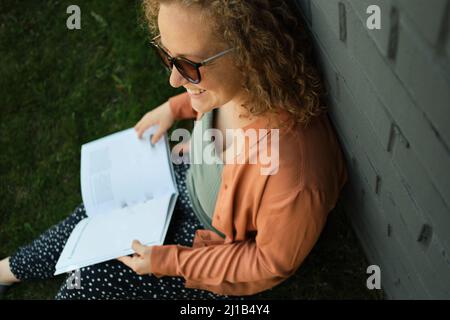 Giovane adulto millennial con capelli ricci sorride e siede sull'erba. Lo studente legge il libro. Stile di vita. La ragazza è calma, tranquilla. La donna indossa occhiali e. Foto Stock