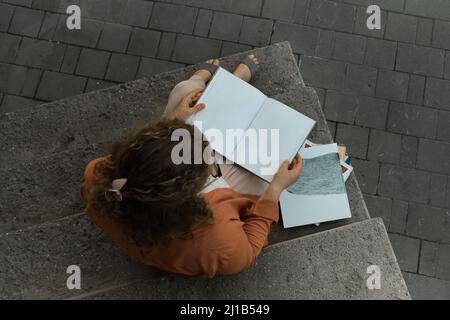 Giovane adulto millenario con capelli ricci siede su gradini. Lo studente legge il libro. Stile di vita in città Foto Stock