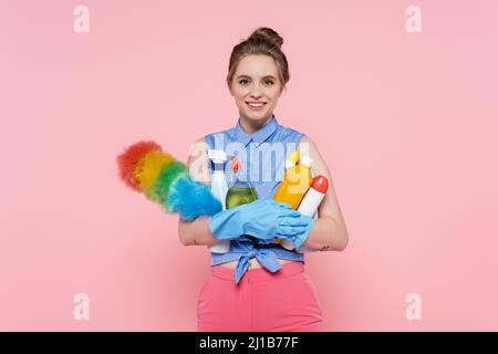 felice giovane donna in guanti di gomma che tengono bottiglie con detergente e spazzolina polvere isolato su rosa Foto Stock