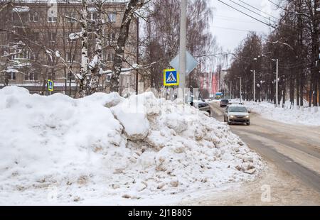 Una grande nevicata sulla strada sullo sfondo di una strada cittadina. Sulla strada si trova neve sporca in cumuli alti. Paesaggio urbano invernale. Nuvoloso giorno d'inverno, luce tenue. Foto Stock
