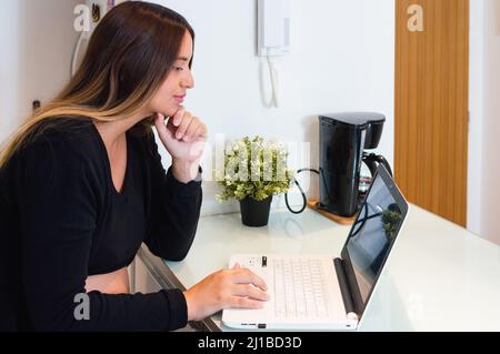 vista laterale donna incinta caucasica che pensa con una mano sul mento a casa guardando lo schermo del laptop, lavorando online con il computer sul banco della cucina. Foto Stock