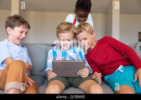 Sorridenti ragazzi della scuola elementare multirazziale che guardano il tablet digitale mentre si siedono sul divano a scuola Foto Stock