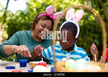 Ragazzo e nonna afro-americana che indossa le orecchie di coniglietto mentre dipinge le uova il giorno di pasqua Foto Stock