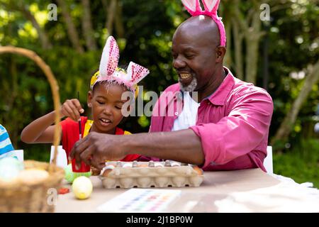 Ragazza e nonno afro-americani che indossano orecchie conigliere mentre dipingi le uova il giorno di pasqua Foto Stock