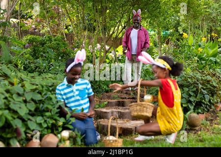 Felici fratelli afroamericani in coniglietto che nascondono le uova di pasqua con il nonno nel cortile Foto Stock