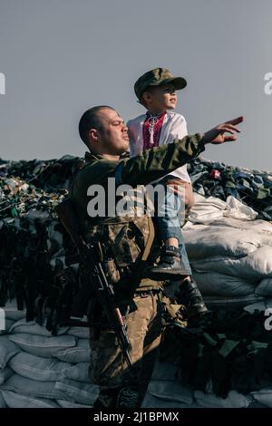 Il guerriero ucraino della difesa territoriale tiene il bambino in camicia ricamata nelle sue mani sul checkpoint. Concetto di invasione militare russa in Ucraina Foto Stock