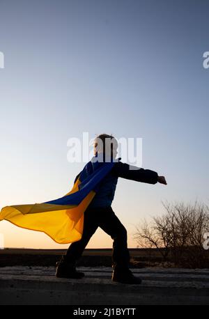 Ritratto di supereroe ragazzo con bandiera Ucraina ondeggiante dietro in posa pronto per la battaglia, mani in pugni. Fermare la guerra. Sostenere l'Ucraina. Infanzia di LIT Foto Stock