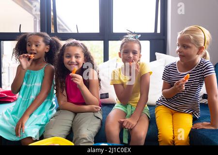 Ritratto di ragazze scolastiche multirazziali elementari sorridenti che mangiano cibo mentre si siedono sul divano a scuola Foto Stock