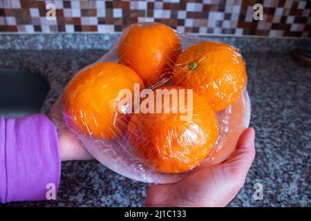 La donna sta tenendo in mano i frutti d'arancia confezionati. Utilizzo di pellicola per alimenti per conservare la frutta in frigorifero. Concetto di conservazione degli alimenti. Foto Stock