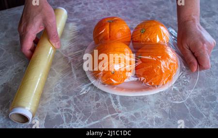 La donna sta tenendo in mano i frutti d'arancia confezionati. Utilizzo di pellicola per alimenti per conservare la frutta in frigorifero. Concetto di conservazione degli alimenti. Foto Stock