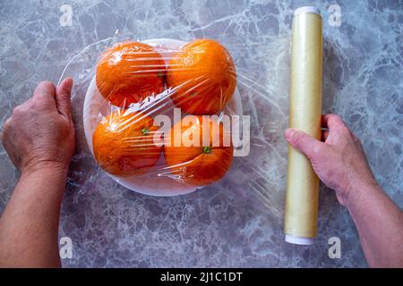 La donna sta tenendo in mano i frutti d'arancia confezionati. Utilizzo di pellicola per alimenti per conservare la frutta in frigorifero. Concetto di conservazione degli alimenti. Foto Stock