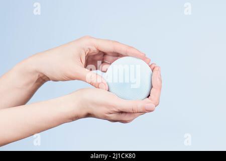 Donna mani che tengono impermeabile silicone pulizia del viso spazzola su sfondo azzurro chiaro. Spazzola moderna per la pulizia del viso, il sollevamento, il massaggio. Vista frontale Foto Stock