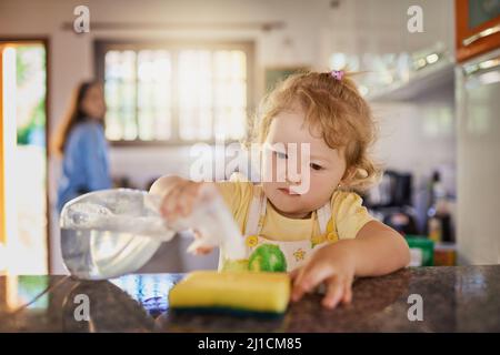 Età le faccende adatte per i bambini sono molto importanti. Shot di una bambina che tiene una bottiglia di detergente detergente con la madre sullo sfondo Foto Stock