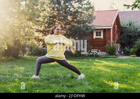 Donna di mezza età adulta facendo esercizi fisici che si allungano per mantenere in forma indossare abiti sportivi sul cortile sul verde erba con casa di campagna in legno Foto Stock