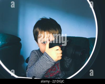 Giovane fotografo che guarda attraverso la fotocamera e gli anelli luminosi, bambino di 2 anni in studio Foto Stock