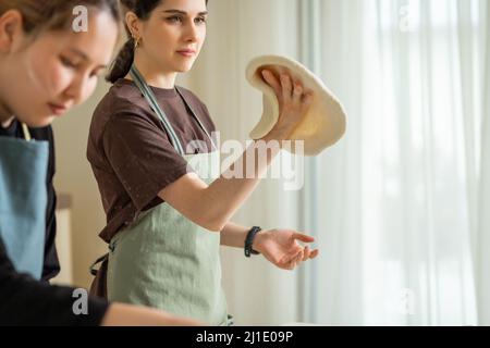 Due giovani cuochi femminili che cucinano la pizza in cucina, uno attorciglia l'impasto arrotolato in mano. Foto Stock