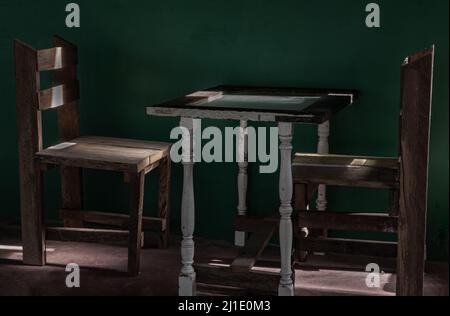 Due sedie di legno e un vecchio tavolo bianco di legno che si trova davanti al muro di cemento verde in una stanza vuota attraverso la quale passa un raggio di luce. Tono scuro, Spac Foto Stock