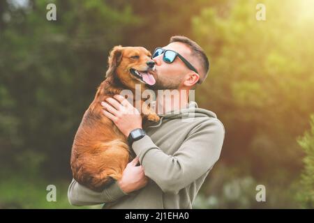 Uomo che tiene il cane tra le braccia e lo bacia all'aperto. Cane e proprietario insieme. Amore per gli animali domestici Foto Stock