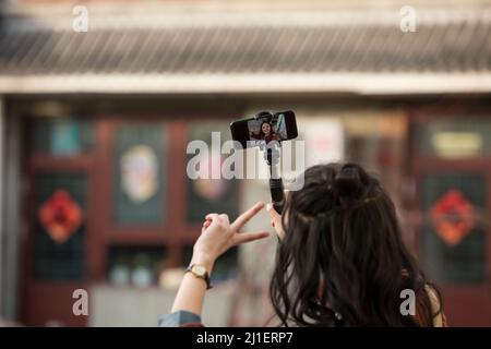 Vista posteriore di capelli ricci lunghi giovane donna cinese che prende selfie con bastone selfie - foto di scorta Foto Stock