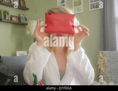 Un primo piano della giovane bella donna che copre il viso con un sacchetto cosmetico rosso, ricevuto come regalo di Natale. Foto Stock