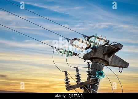 Palo elettrico in calcestruzzo con isolatori in vetro su cavi elettrici, su cielo giallo e blu al tramonto Foto Stock