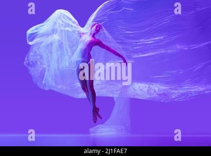 Ritratto di bella donna flessibile, ballerina ballando con panno su sfondo viola studio in neon. Moda, stile, arte, bellezza Foto Stock