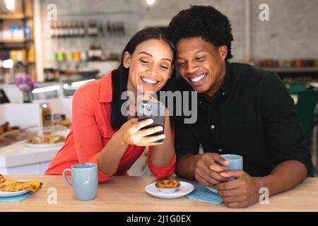 Felice giovane coppia multirazziale condividere smartphone mentre si siede con caffè insieme in caffè. Inalterato, caffè cultura, persone, amore, tecnologia e li Foto Stock
