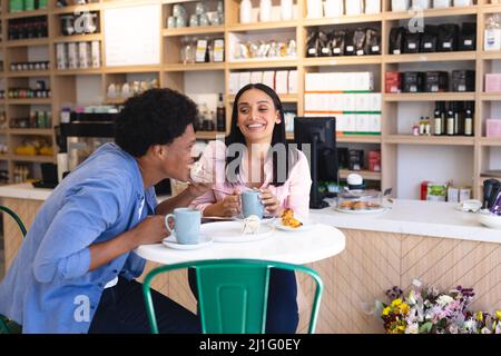 Allegra coppia di giovani multirazziali che gustano caffè e snack al tavolo in un caffè. Intatta, cultura del caffè, persone, amore e stile di vita concetto. Foto Stock