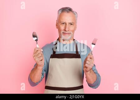 Foto di divertente affascinante uomo in pensione indossare camicia grigia grembiule tenere forcella cucchiaio isolato rosa sfondo di colore Foto Stock