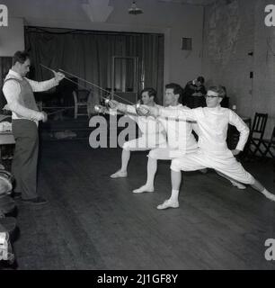 1960s, storico, tre studenti di sesso maschile che indossano tradizionali calzoni, giacche e pilmsols in una classe di scherma, con istruttore, Università di Oxford, Inghilterra, Regno Unito. Essi praticano la spinta dei fencer, un movimento chiave nello sport, che è una spinta attaccante fatta con un piede in avanti e la gamba posteriore dritta e il braccio della spada allungato in avanti. Foto Stock